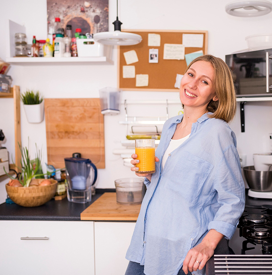 A woman in her kitchen holds a glass of orange juice, smiling as she enjoys a refreshing drink.