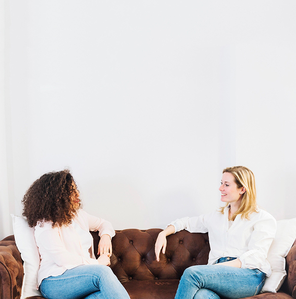 Two women are seated on a brown couch, engaged in conversation and smiling at each other.