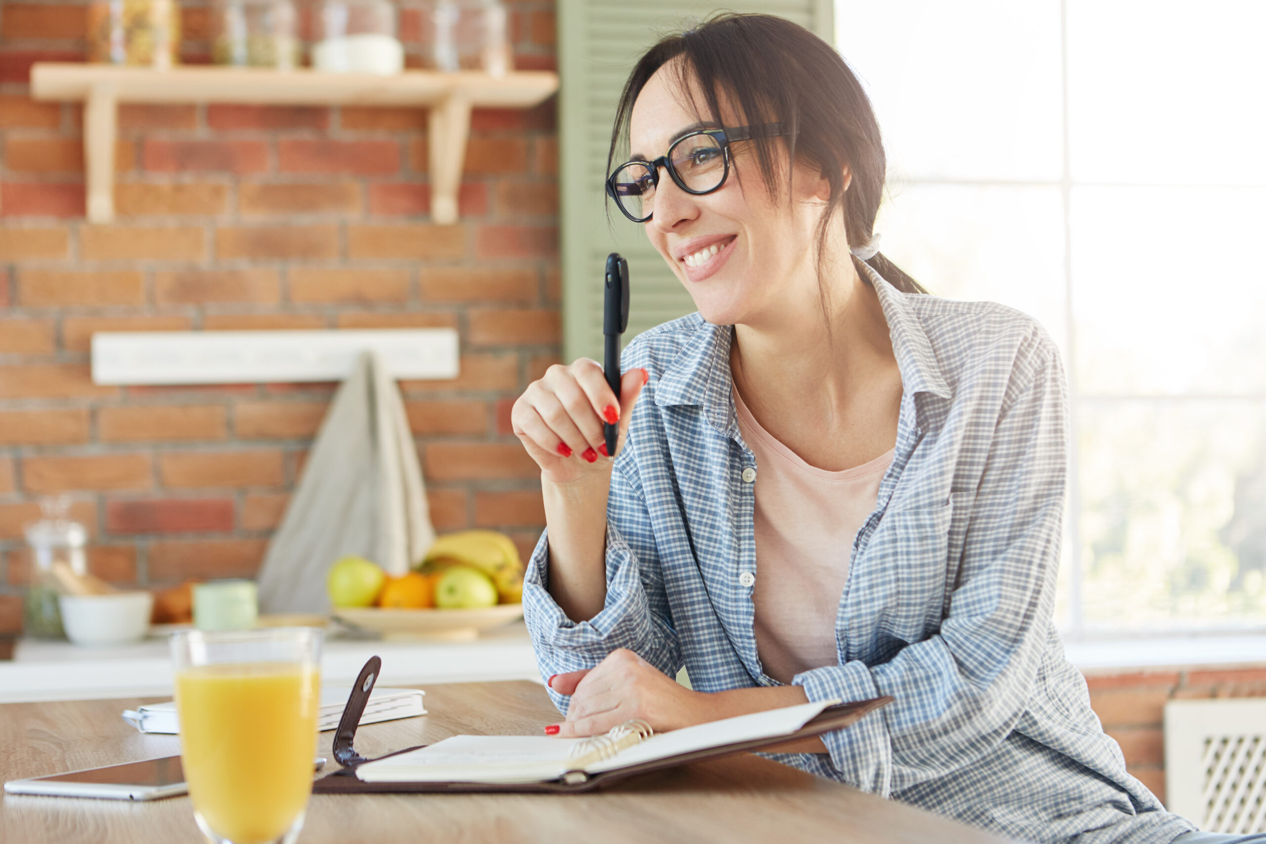 Happy woman with appealing appearance, going to organize party, makes list of invited friends, sits in kitchen, drinks orange juice, holds pen, writes in notepad salad recipe. Home atmosphere